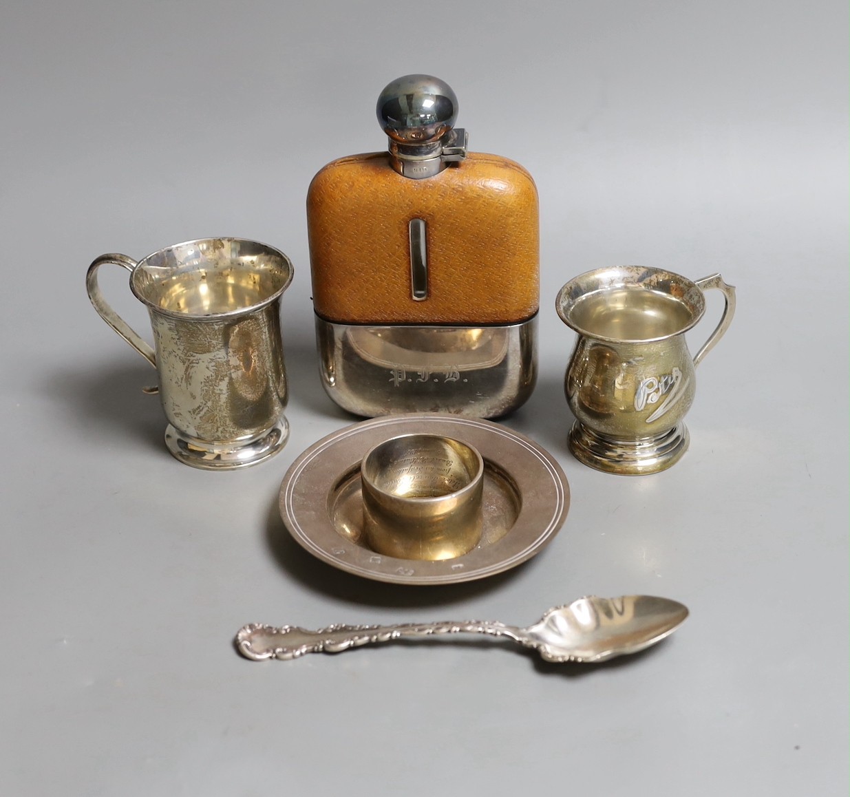 Two small George V christening mugs, a small modern silver armada dish, a silver napkin ring, ornate silver spoon and a silver plate and leather mounted glass hip flask.